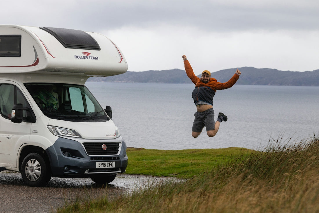 A man jumping for joy beside a white motorhome near a coastal cliff